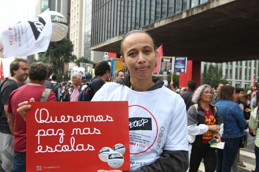 Professora vestida com a camisa da Apeoesp segura nas mãos um cartaz em que pede "paz nas escolas"