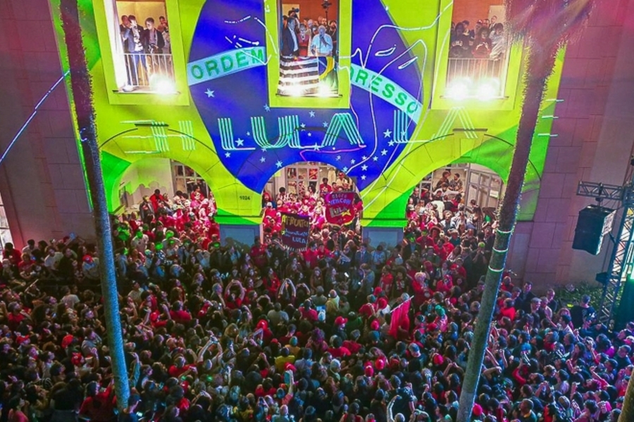Foto da sacada da entrada da PUC, em que Lula e Haddad acenam para uma multidão.