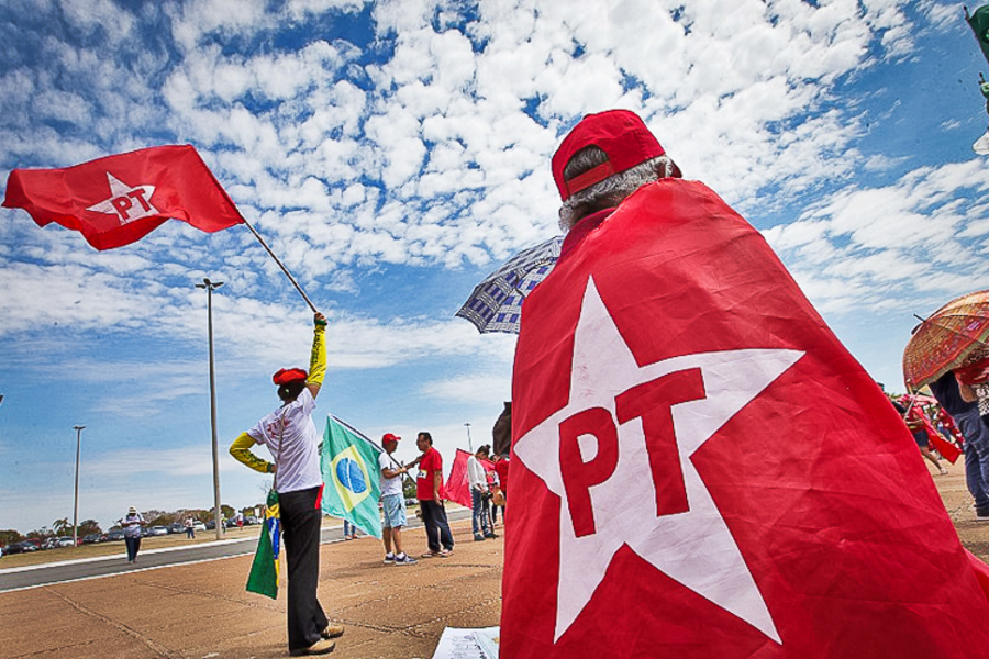 Homem caminha com a bandeira do PT enrolada nas costas, enquanto outro tremula outra bandeira do Partido