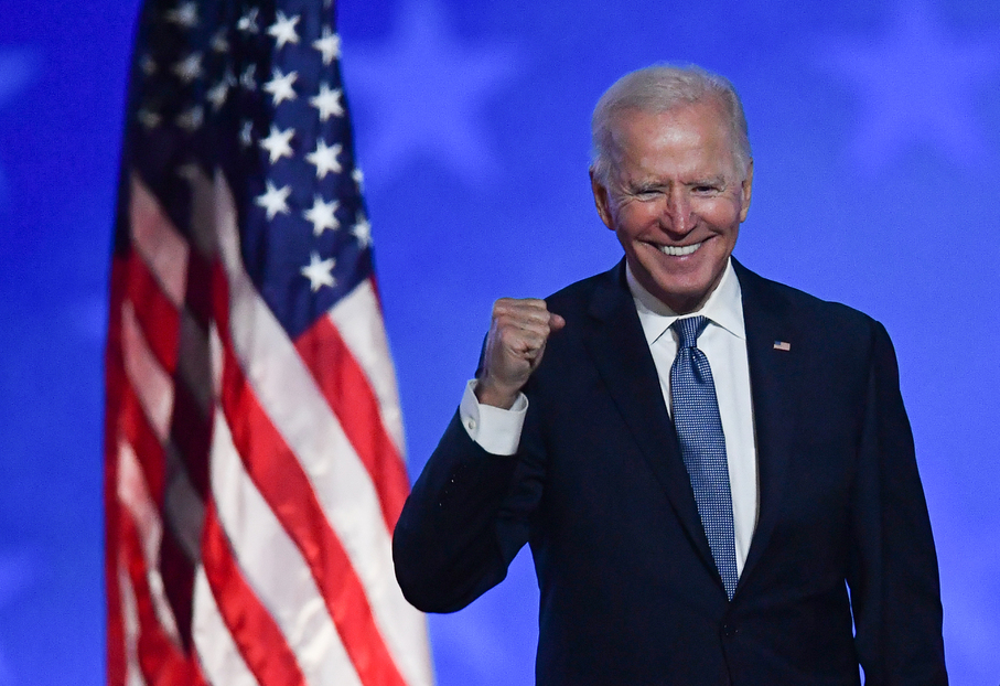 Joe Biden com o braço meio estendido e punhos fechados sorri com a bandeira norteamericana atrás