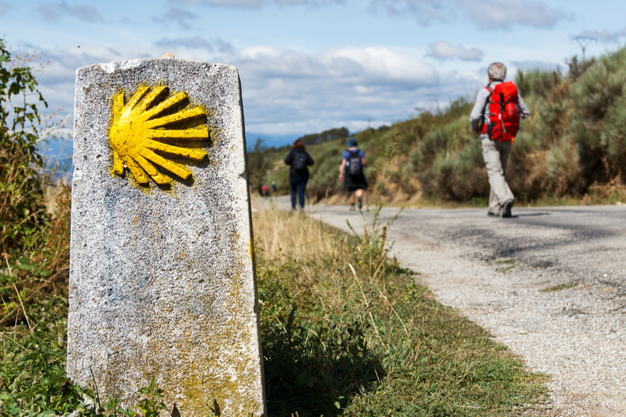 Foto mostra um totem no caminho de Santiago de Compostela. Ao fundo, uma pessoa caminha.