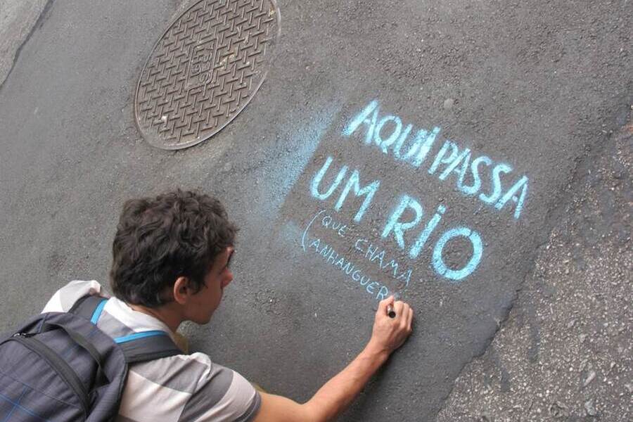 Criança com mochila nas costas escreve no chão de uma rua: "Aqui passa um rio".