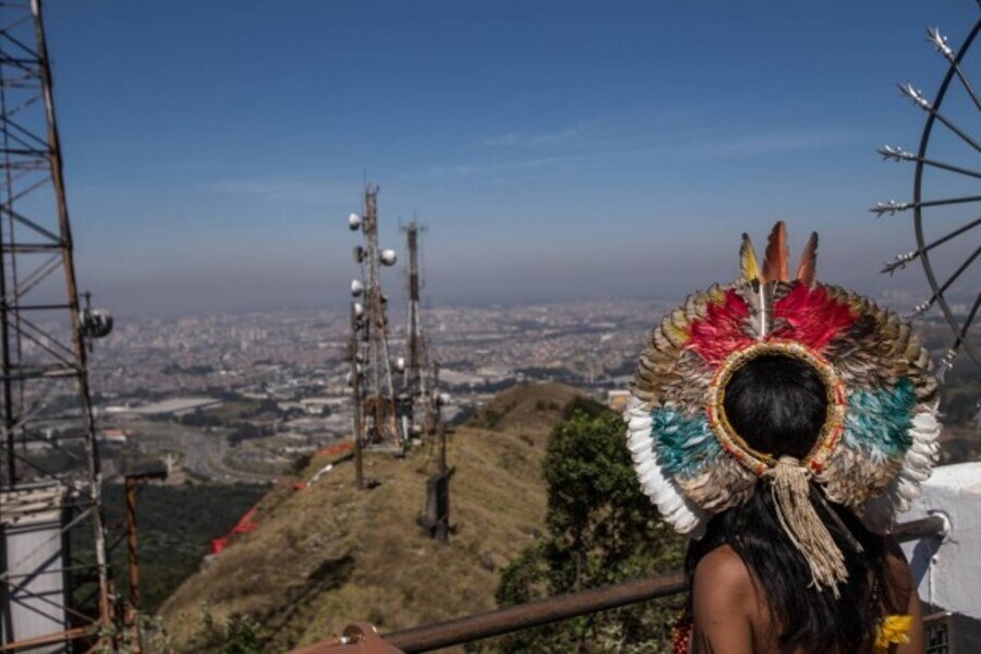 Indígena com seu cocar observa do alto, a cidade de São Paulo