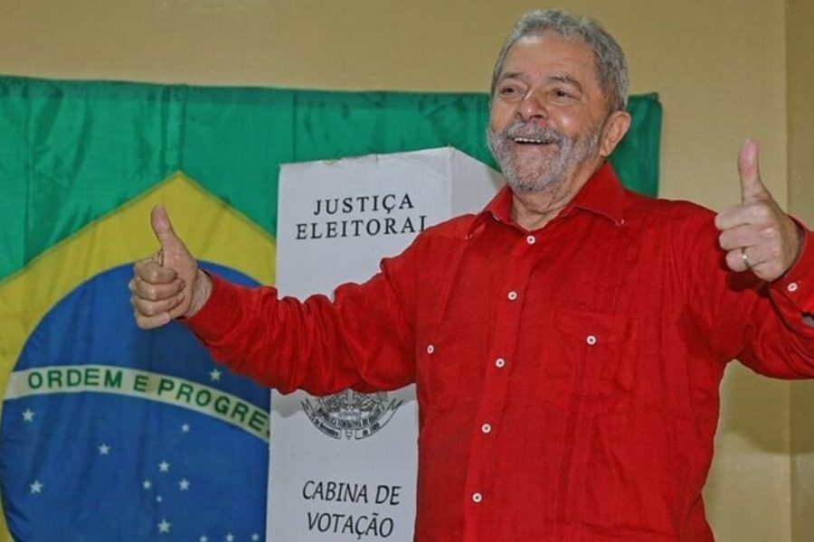 Ex-presidente Lula, de camisa vermelha, sorri e abre os braços fazendo sinal de positivo, após votar. Ao fundo, a bandeira Brasileira e uma urna eleitoral