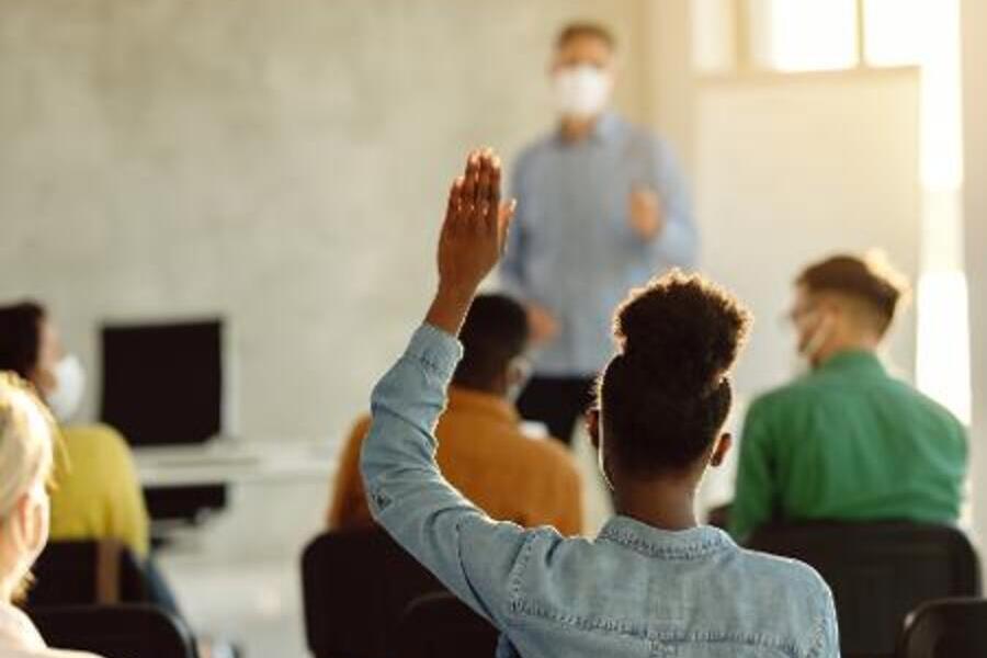 Aluna negra ergue a mão, pedindo a palavra, ao professor dentro da sala de aula