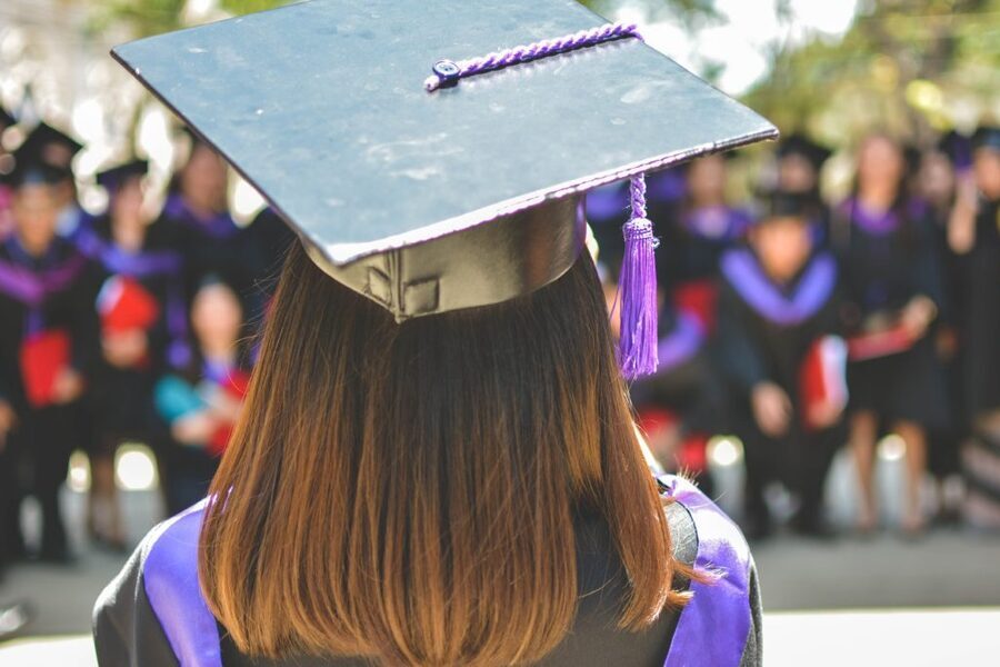 Uma estudante olha de frente para seus colegas. Ela está com a indumentária na cabeça que indica que ela é uma recém formada