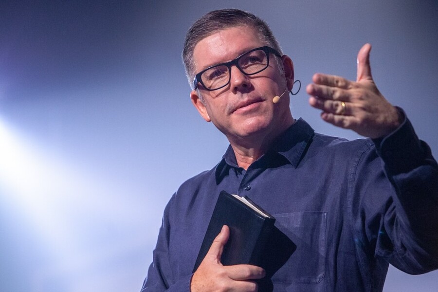 Foto do Pastor Ed Rene Kivitz ergue o braço esquerdo, enquanto segura sob o peito, um exemplar da bíblia.