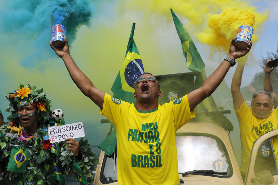 Militante bolsonarista vestido com uma camisa amarela e segurando com ambas as mãos e de braços abertos, sinalizadores que soltam fumaça colorida