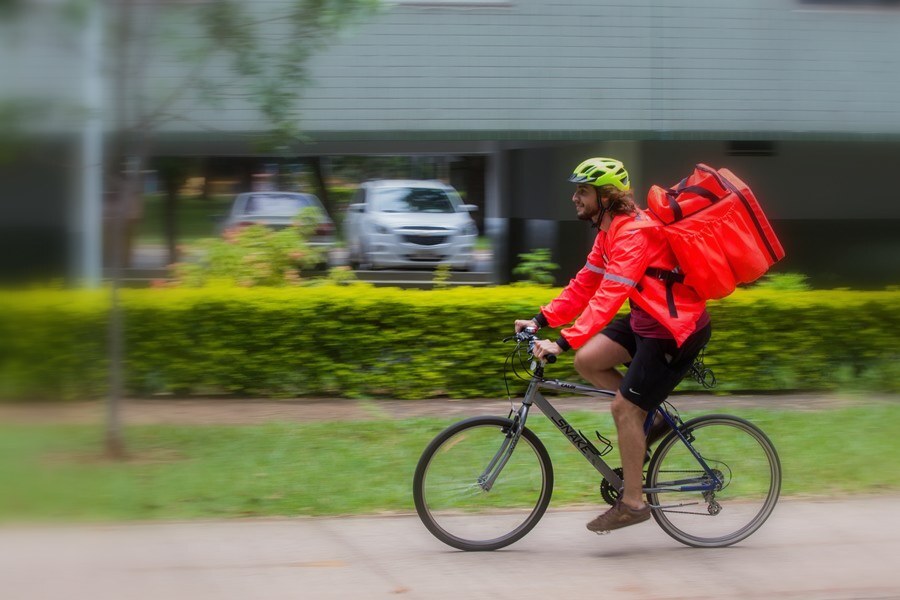 Entregador por aplicativo pilotando sua bicicleta, indo da direita para a esquerda